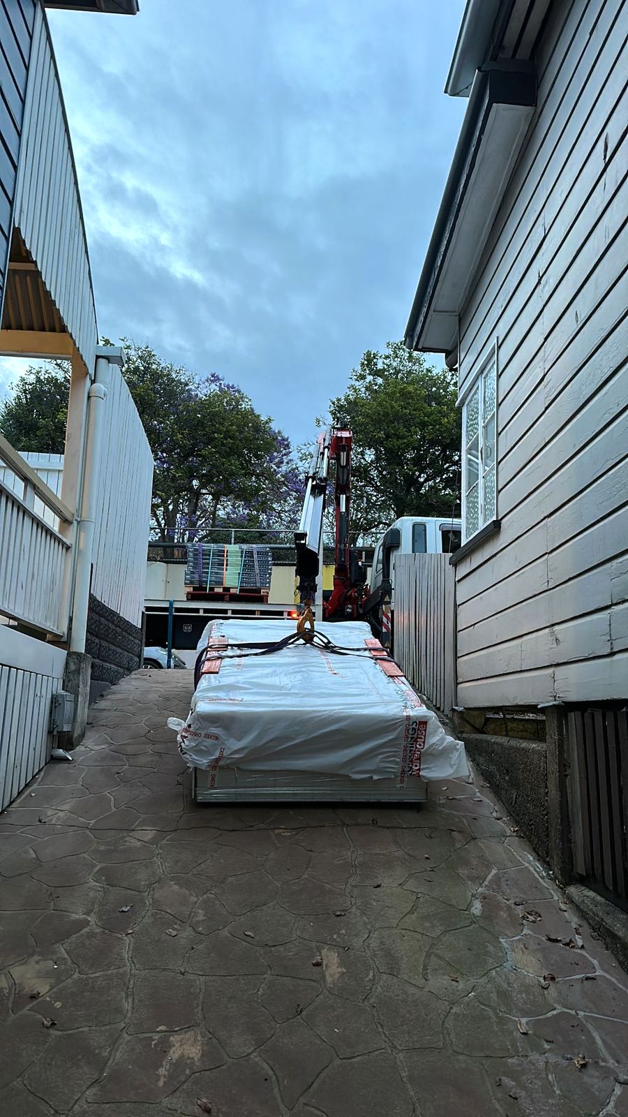 Dingo crane truck lifting a wrapped pack of building materials down a narrow side path between two houses at dusk